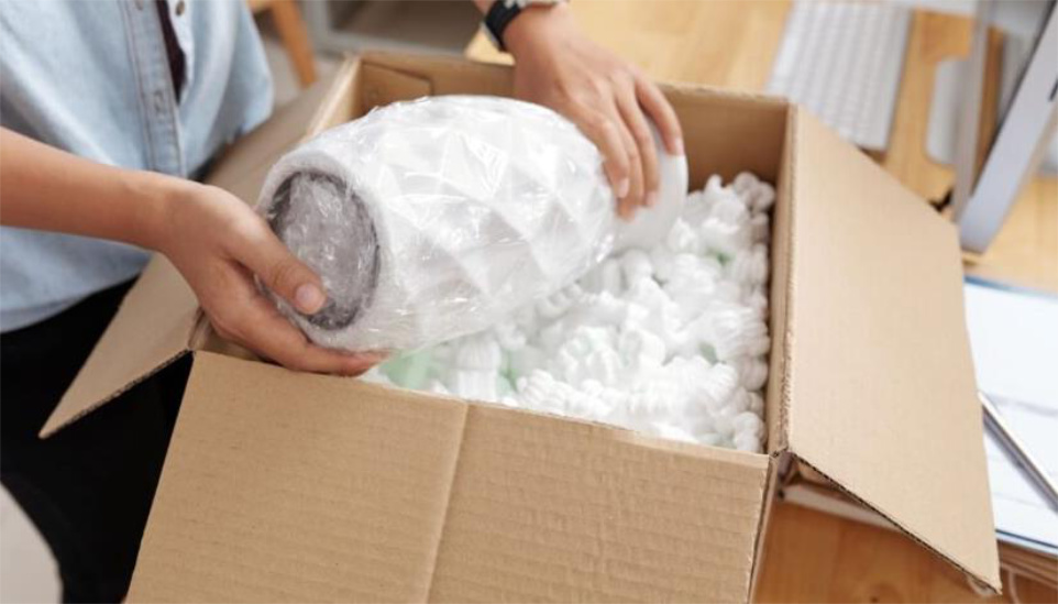 Person placing a bubble-wrapped item in a box with packing peanuts